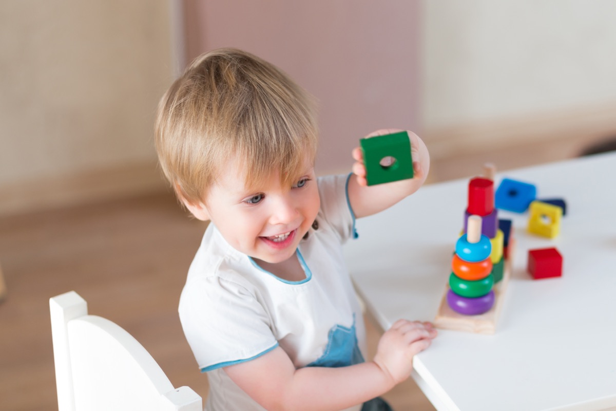 A smiling toddler at a small table stacking wooden shape blocks